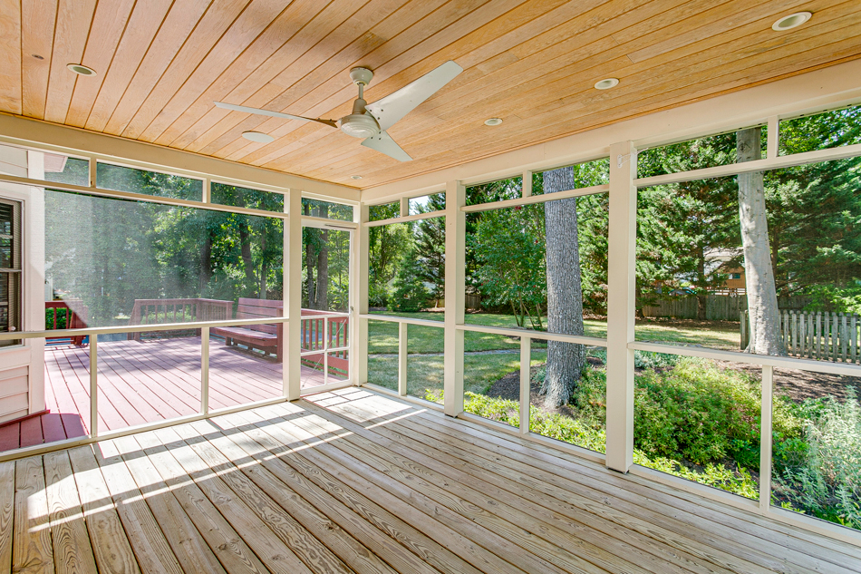 Spacious-Screened-In-Porch-With-Ceiling-Fan-And-Deck-Overlooking-Green-Backyard-2163703117 a spacious screened-in porch equipped with a ceiling fan, wooden decking, and large screened windows providing a view of a lush green backyard. The area is designed for both comfort and enjoying the outdoors while keeping pests at bay.